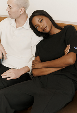A man and a woman sitting side by side indoors. The woman wears a black short-sleeve top and black pants, and the man wears a white polo shirt and dark pants.