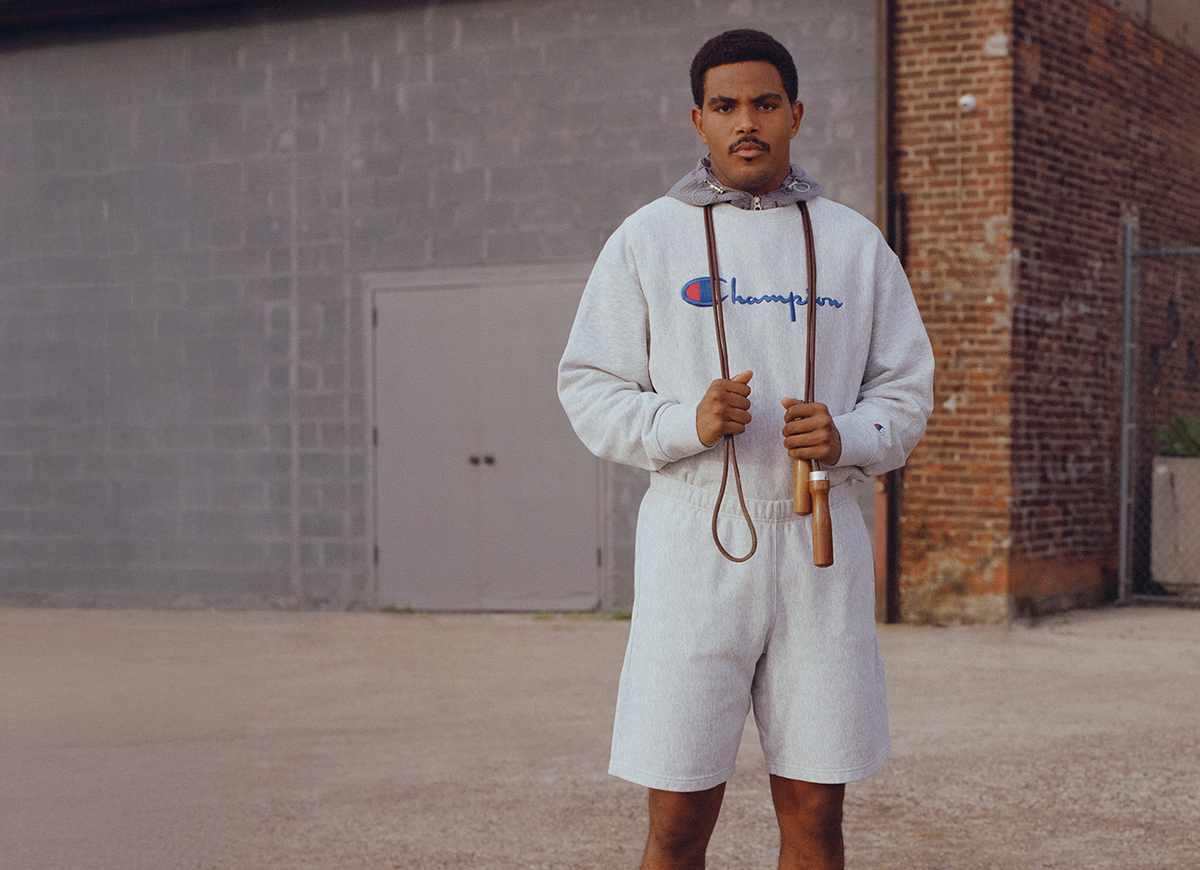 A man wearing a light gray Champion hoodie and matching shorts stands outdoors holding a wooden jump rope in front of a gray brick wall.