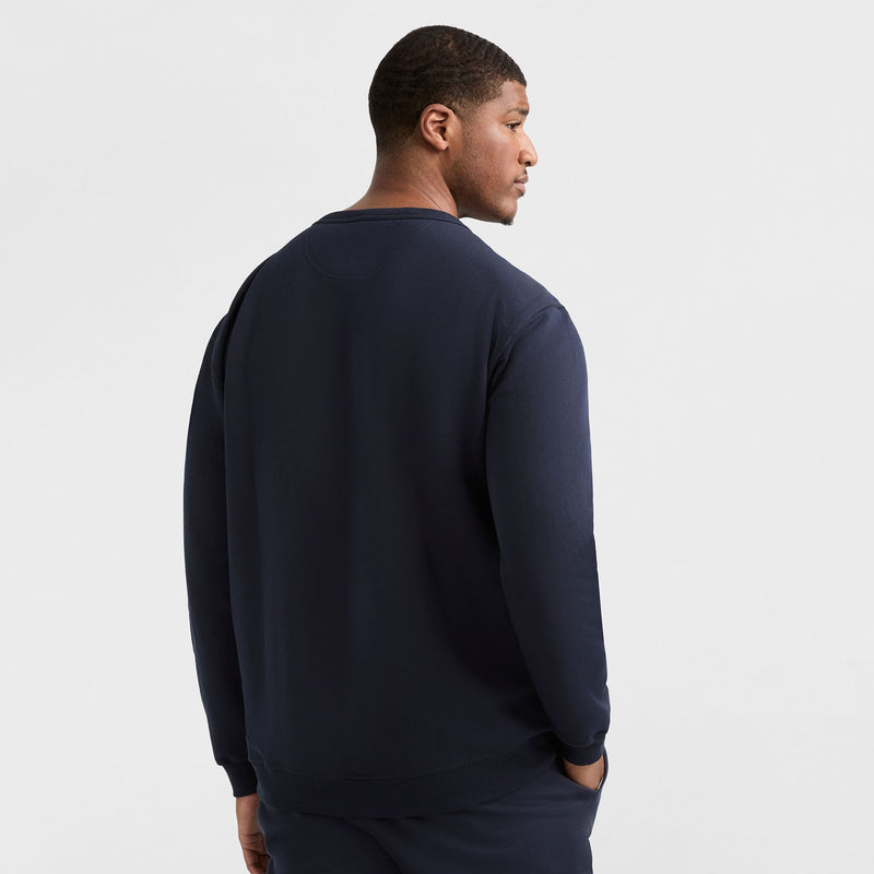 A man faces away from the camera, wearing a dark navy Champion Powerblend Crewneck Sweatshirt, C Logo - Big & Tall and matching pants, set against a plain white background.