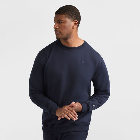 A man wears a navy Champion Powerblend Crewneck Sweatshirt, C Logo - Big & Tall, and matching pants, standing against a white background with his hands loosely clasped, looking to his right.