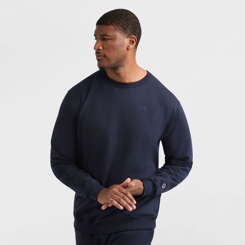 A man wears a navy Champion Powerblend Crewneck Sweatshirt, C Logo - Big & Tall, and matching pants, standing against a white background with his hands loosely clasped, looking to his right.