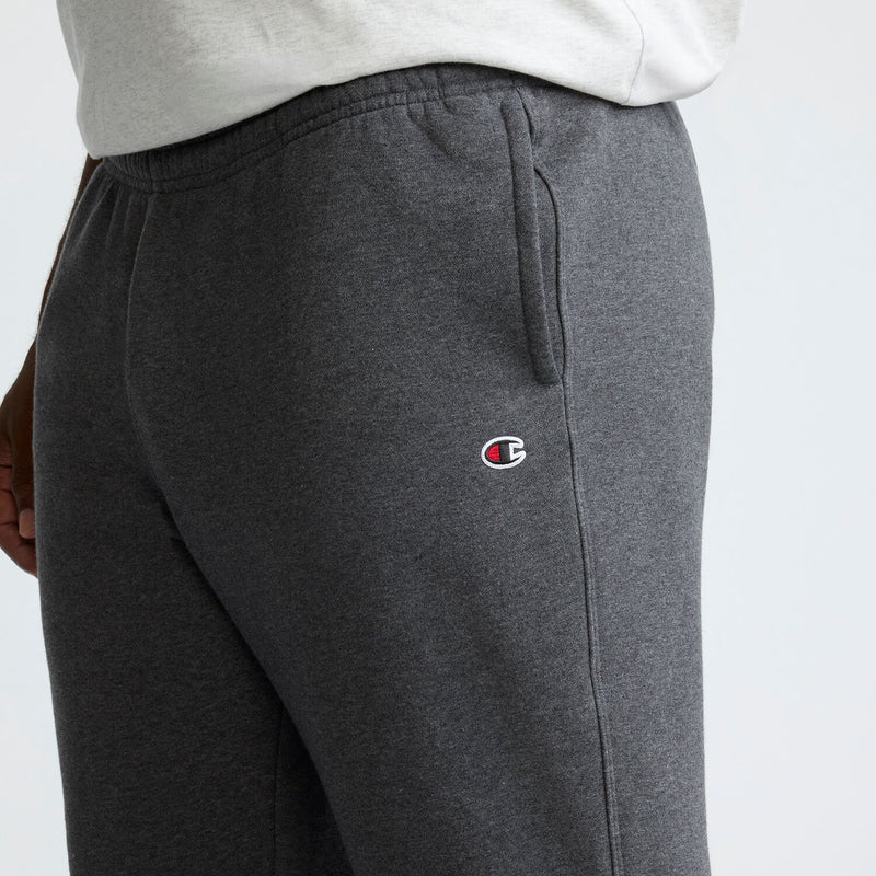 A person wearing dark gray Champion Powerblend Sweatpants, Cinched Bottom, C Logo - Big & Tall, with a small Champion logo on the upper thigh and a light gray T-shirt. Only the midsection and upper legs are visible.