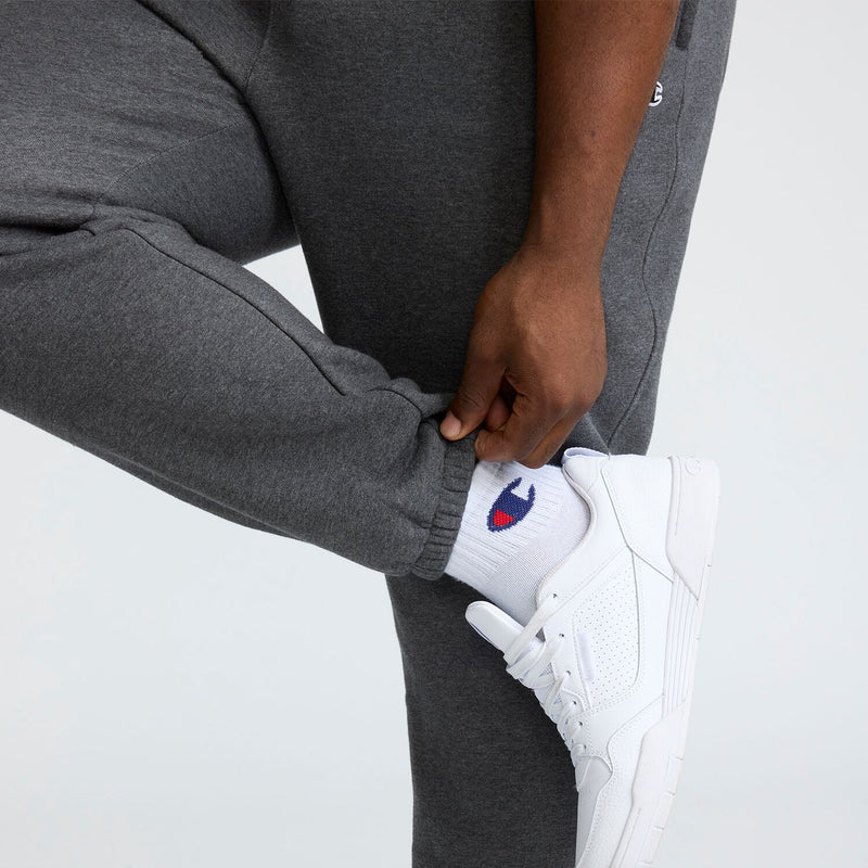 A person in Champion Powerblend Sweatpants, Cinched Bottom, C Logo - Big & Tall adjusts the cuff over white socks with a blue and red logo, paired with white sneakers against a plain light background.