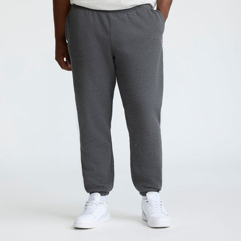 A person stands with hands in pockets, wearing Champion Powerblend Sweatpants, Cinched Bottom, C Logo - Big & Tall in gray and white sneakers. The image is cropped from shoulders down against a plain light background.