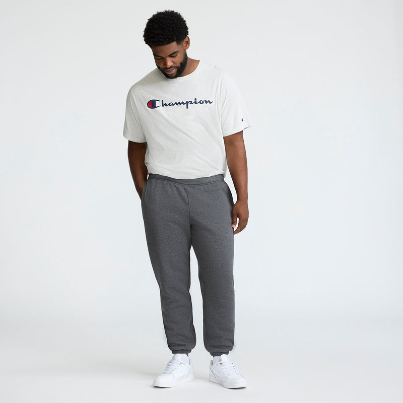 A man stands against a white background, wearing a light gray Champion t-shirt and Champion Powerblend Sweatpants, Cinched Bottom, C Logo - Big & Tall in dark gray with white sneakers. He looks down, smiling with hands in his pockets.