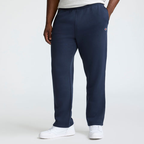 A person wearing Champion Powerblend Sweatpants, Open Hem, C Logo - Big & Tall in navy blue, a light gray T-shirt, and white sneakers stands against a plain background. Only the lower half of the body is visible.