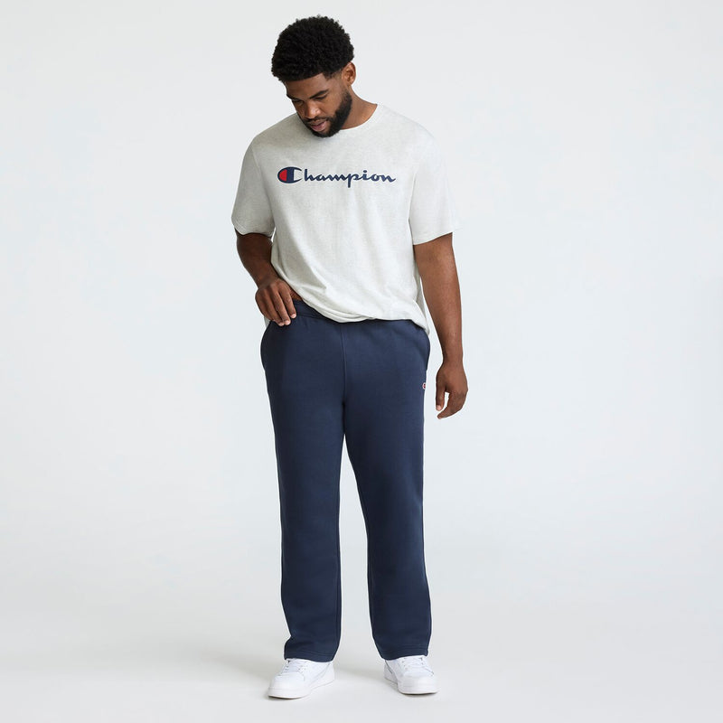 A man stands against a plain light background wearing a light gray t-shirt, Champion Powerblend Sweatpants, Open Hem, C Logo - Big & Tall in navy blue, and white sneakers. He looks down with one hand in his pocket.