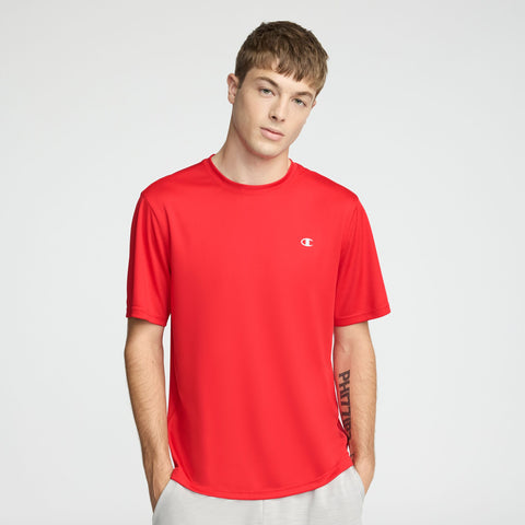 Young man with short brown hair wearing a bright red Champion Sport T-Shirt with Reflective C Logo and light gray pants, standing with hands in pockets against a plain light background.
