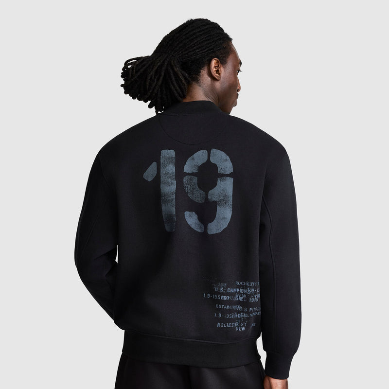A person with long hair in a black Champion Fleece Baseball Jacket stands facing away, displaying a large faded 19 and smaller printed text on the back, evoking a vintage athletic style reminiscent of a classic varsity jacket.