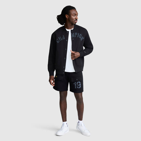 A man stands against a plain light gray background, looking to his left. He is wearing a black Champion Fleece Baseball Jacket, black shorts featuring the number 19 for a vintage athletic touch, a white T-shirt, and white sneakers.