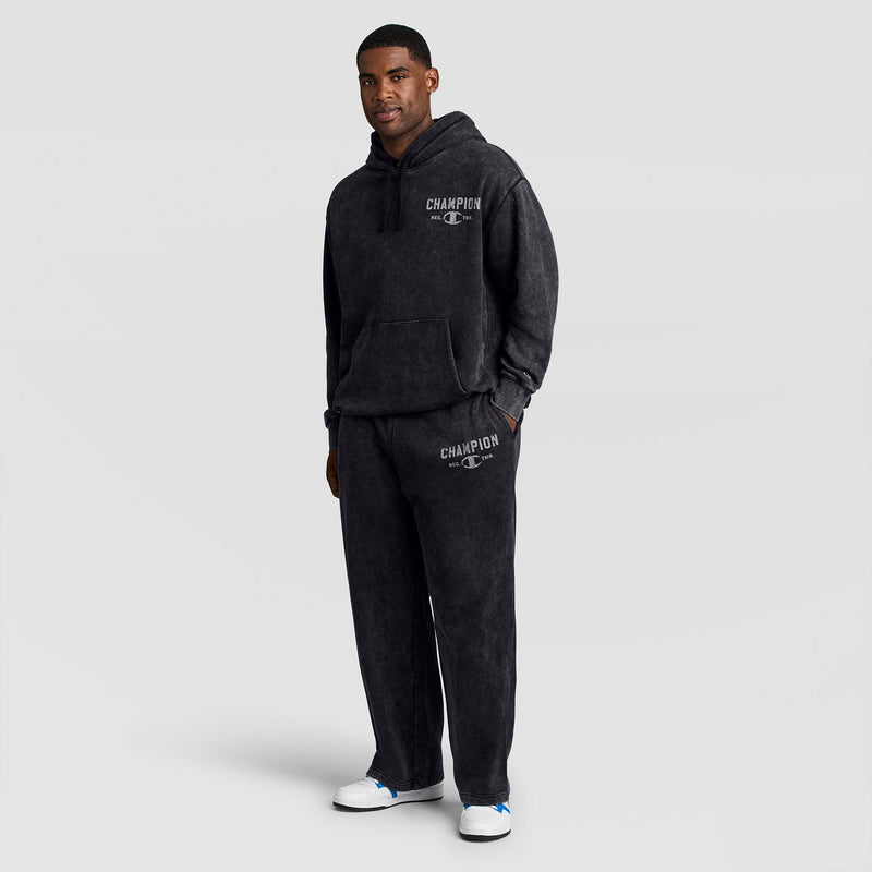 A man wearing a black mineral wash hooded sweatshirt and matching Champion Mineral Wash Open Hem Pant, each featuring a logo on the chest and thigh.
