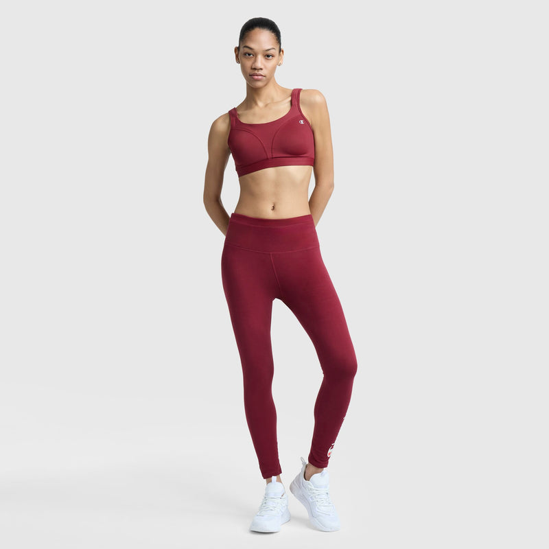 A woman stands against a white background, wearing the Champion Spot Comfort High-Impact Sports Bra with C Logo and matching leggings, paired with white sneakers. Her hands are on her hips and she looks straight ahead with a neutral expression.
