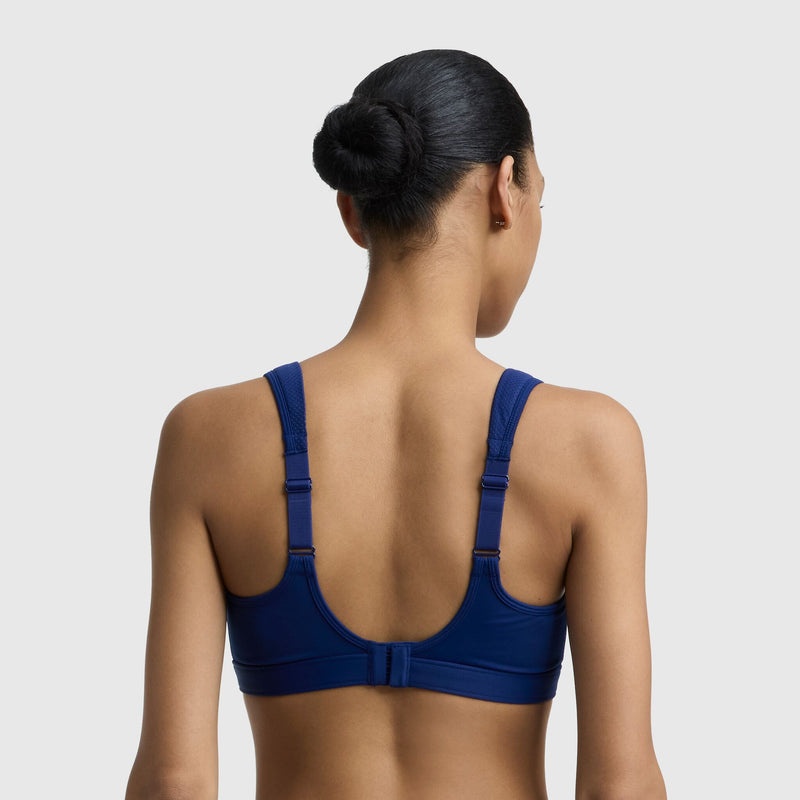 A woman with dark hair in a bun is seen from the back wearing the Champion Spot Comfort High-Impact Sports Bra, C Logo, in blue with thick adjustable straps and a clasp closure. The plain background highlights its maximum support design.
