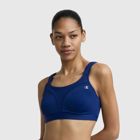 A woman with pulled-back hair wears the Champion Spot Comfort High-Impact Sports Bra with C Logo in blue, standing against a plain light gray background.