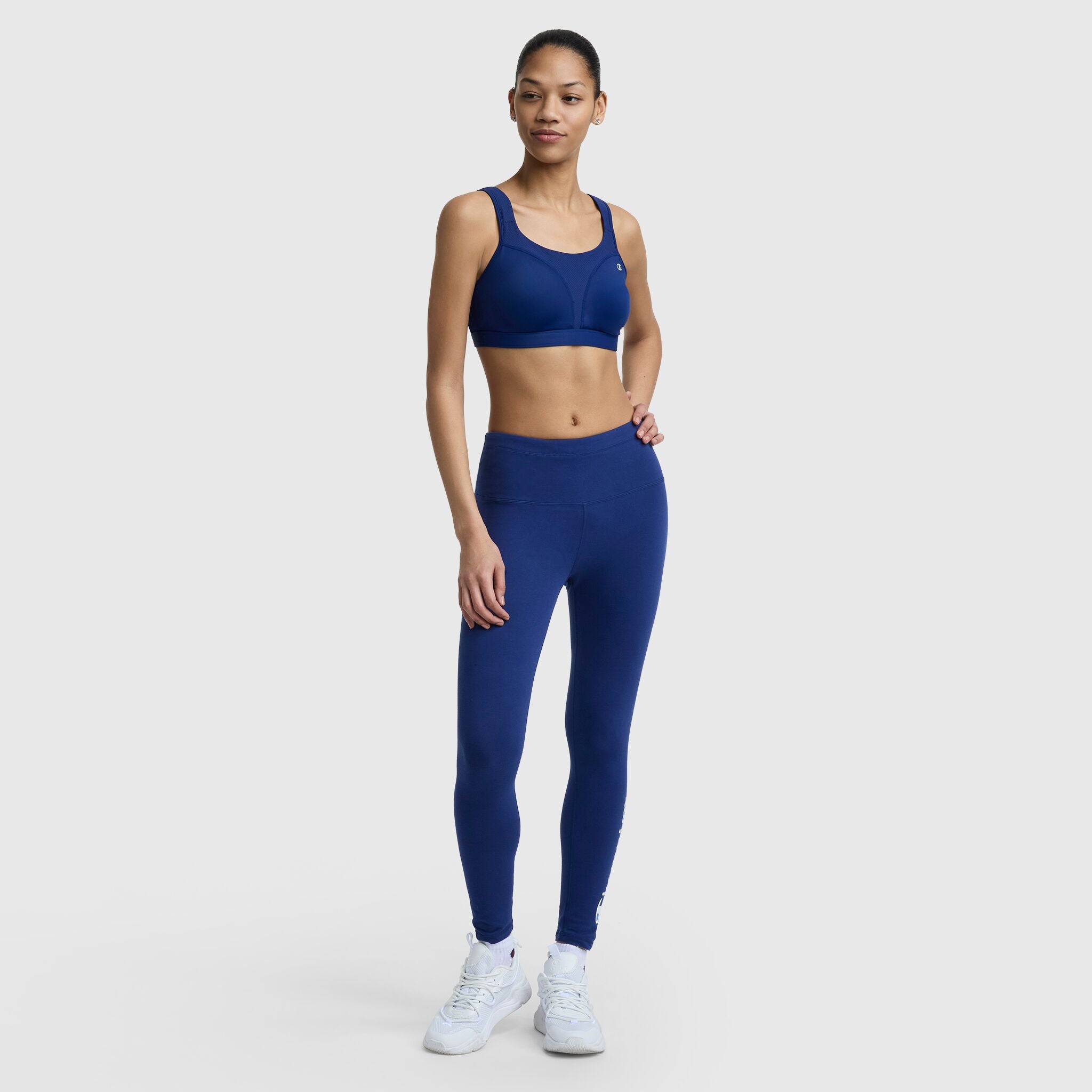 A woman stands confidently in a Champion Spot Comfort High-Impact Sports Bra, C Logo, and matching leggings with white sneakers against a plain light background.