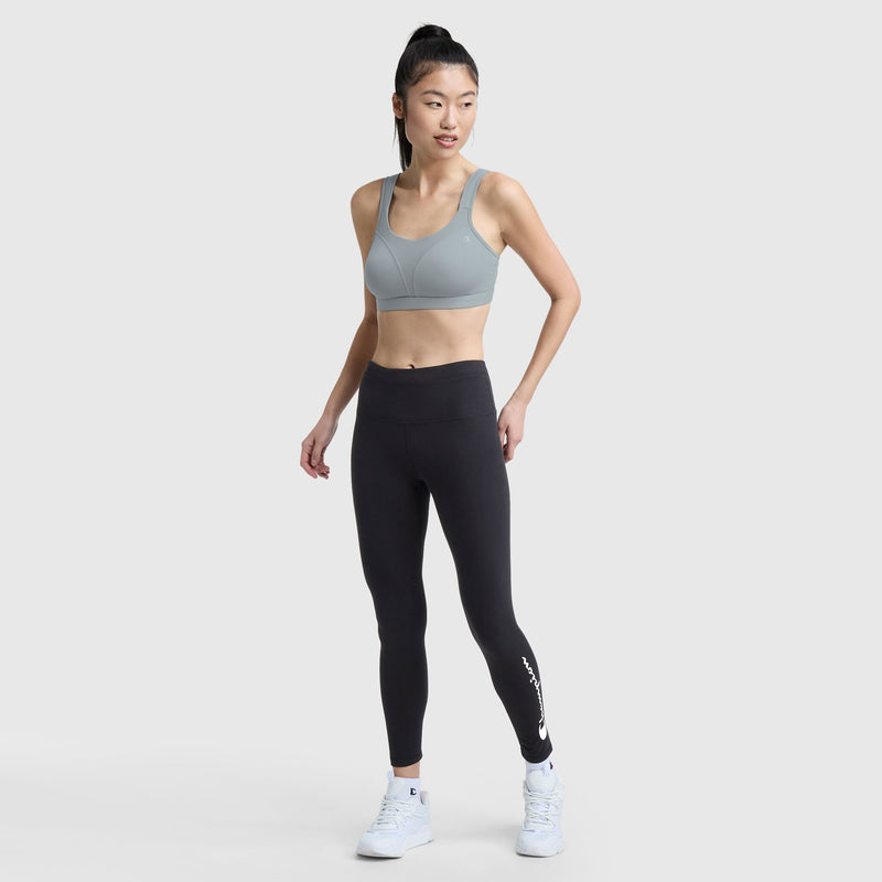 A woman stands in a relaxed pose, wearing the Champion Spot Comfort High-Impact Sports Bra with C Logo, paired with high-waisted black leggings featuring white text near the ankle, and white sneakers against a plain light gray background.
