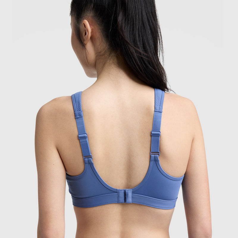 A woman with long dark hair in a ponytail is shown from the back, wearing the Champion Spot Comfort High-Impact Sports Bra, C Logo. The blue bra features adjustable straps and a hook-and-eye closure against a plain light background.