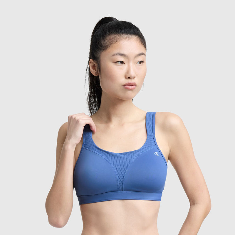 A woman with a high ponytail wears the Champion Spot Comfort High-Impact Sports Bra, C Logo, in blue. She stands against a plain light grey background and looks to the side, holding one strap of her sports bra with her right hand.