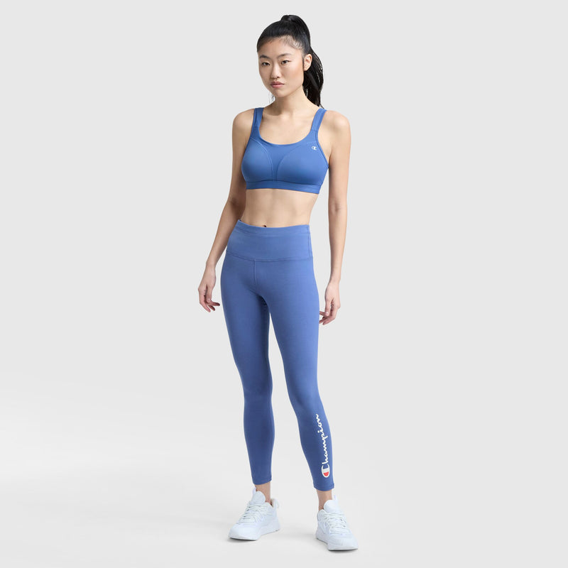 A woman with long black hair in a high ponytail wears Champion's Spot Comfort High-Impact Sports Bra, C Logo in blue and matching high-waisted leggings with a Champion logo, paired with white sneakers, standing against a plain light background.