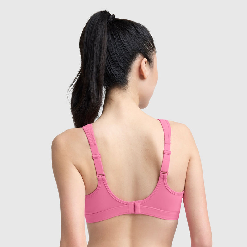 A woman with long dark hair in a ponytail is shown from the back, wearing the Champion Spot Comfort High-Impact Sports Bra, C Logo, with wide straps and hook-and-eye closure. The plain light gray background highlights its maximum support design.