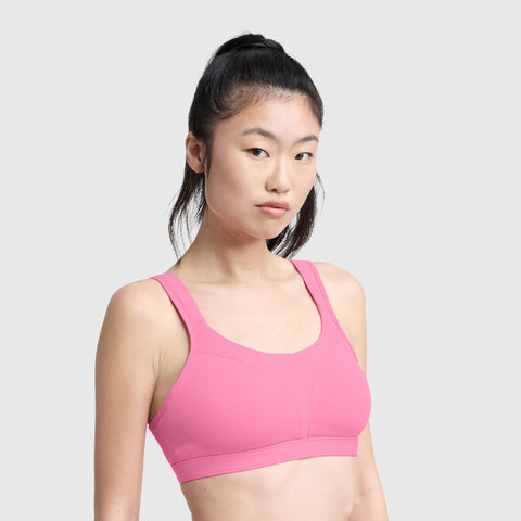 A woman with long black hair in a ponytail wears the Champion Spot Comfort High-Impact Sports Bra, C Logo, in pink. She stands against a light gray background, looking at the camera with a neutral expression.