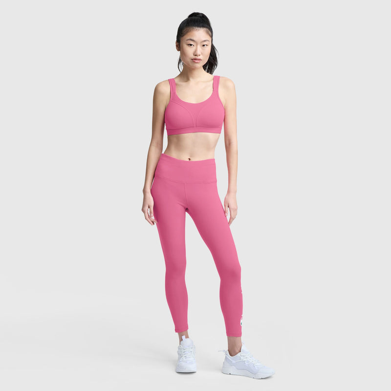 A woman poses against a plain light gray background wearing the Champion Spot Comfort High-Impact Sports Bra, C Logo in pink, matching leggings, and white sneakers.