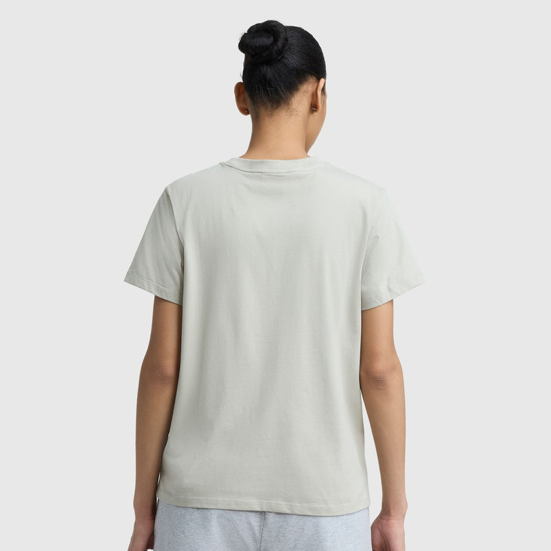 A person with dark hair in a bun is seen from behind, wearing a Champion Classic T-Shirt with tonal left chest script in light beige and light gray shorts against a plain white background.