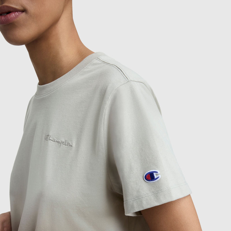 A person wears a light beige women's Champion Classic T-Shirt, Tonal Left Chest Script, featuring an embroidered logo on the chest and a small sleeve patch, set against a plain light background.