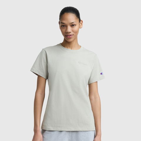 A woman with dark hair tied back models the Champion Classic T-Shirt, Tonal Left Chest Script and gray pants against a plain white background. The light gray tee features subtle logos on the chest and sleeve.