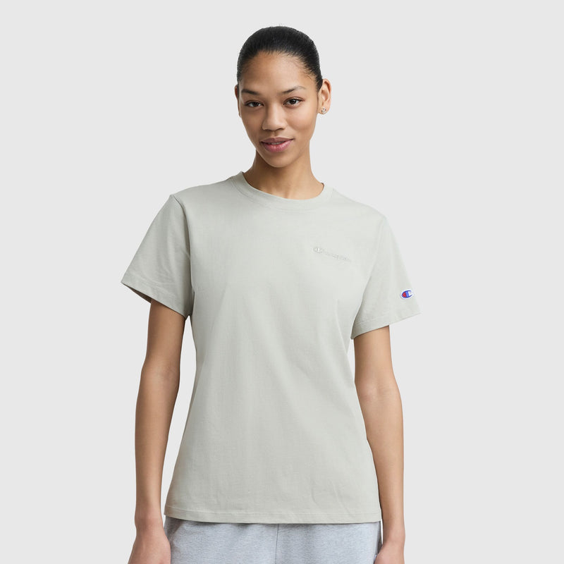 A woman with dark hair tied back models the Champion Classic T-Shirt, Tonal Left Chest Script and gray pants against a plain white background. The light gray tee features subtle logos on the chest and sleeve.