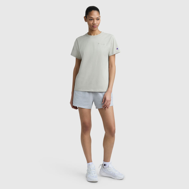 A woman stands against a plain light background, wearing a light gray Champion Classic T-Shirt with Tonal Left Chest Script, gray shorts, white socks, and white sneakers. She looks slightly to the side with a relaxed expression.