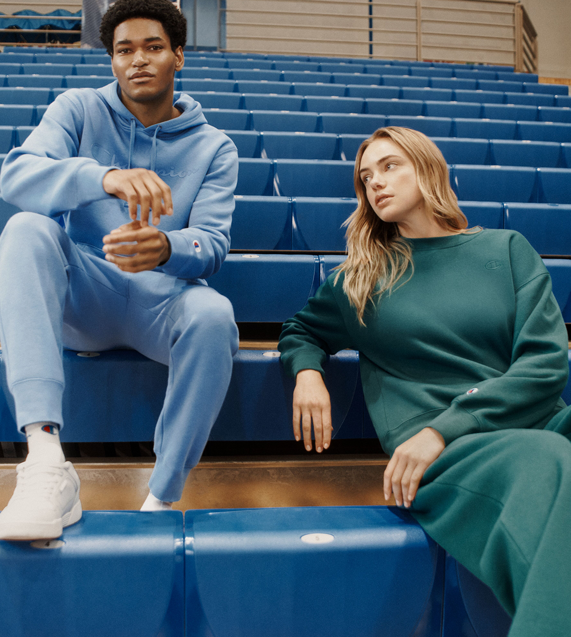Two models sitting on blue stadium seating in an indoor arena, wearing matching oversized sweatshirt and jogger sets in light blue and dark green. The joggers feature a small embroidered Champion 'C' logo on the chest.
