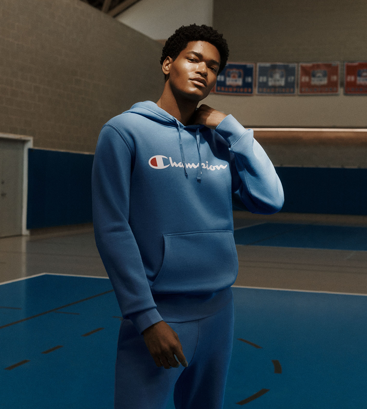 A man wearing a blue Champion hoodie and matching sweatpants stands on an indoor basketball court. The hoodie features the white Champion logo across the chest.