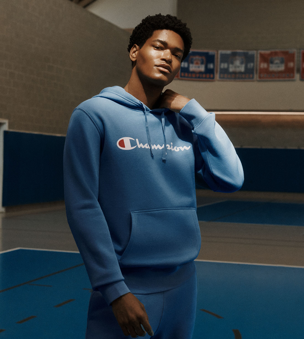 A man wearing a blue Champion hoodie and matching sweatpants stands in a gymnasium with a blue court and wall visible behind him. The hoodie has the Champion logo across the chest in white with a red and blue C.