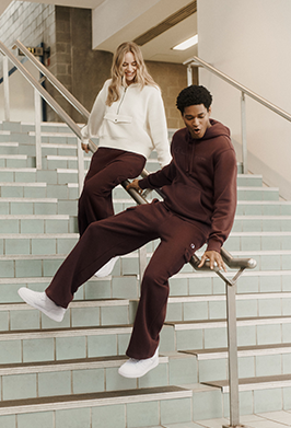 A man and a woman modeling matching burgundy sweatpants and hoodies on an indoor staircase. The man sits on the handrail wearing a burgundy hoodie and matching sweatpants with white sneakers, while the woman stands behind him in a white hoodie and matching burgundy pants.