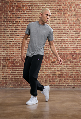 Man in gray t-shirt and black pants walking against a brick wall.