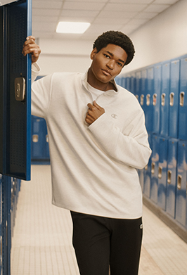A young man wearing a white long-sleeve quarter-zip pullover and black sweatpants stands in a school locker room next to blue lockers.