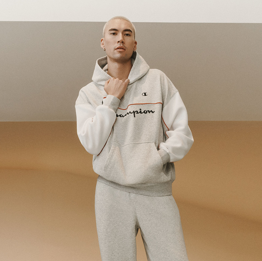 A man modeling a light gray Champion hoodie and matching sweatpants set in an indoor staircase area. The hoodie features a white sleeve, a light gray body, and a thin orange accent line above the Champion logo, which is embroidered in black.