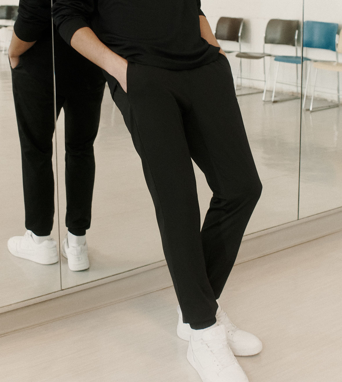 A person wearing black slim-fit pants and a black top, standing in a bright studio with mirrored walls and white sneakers visible.