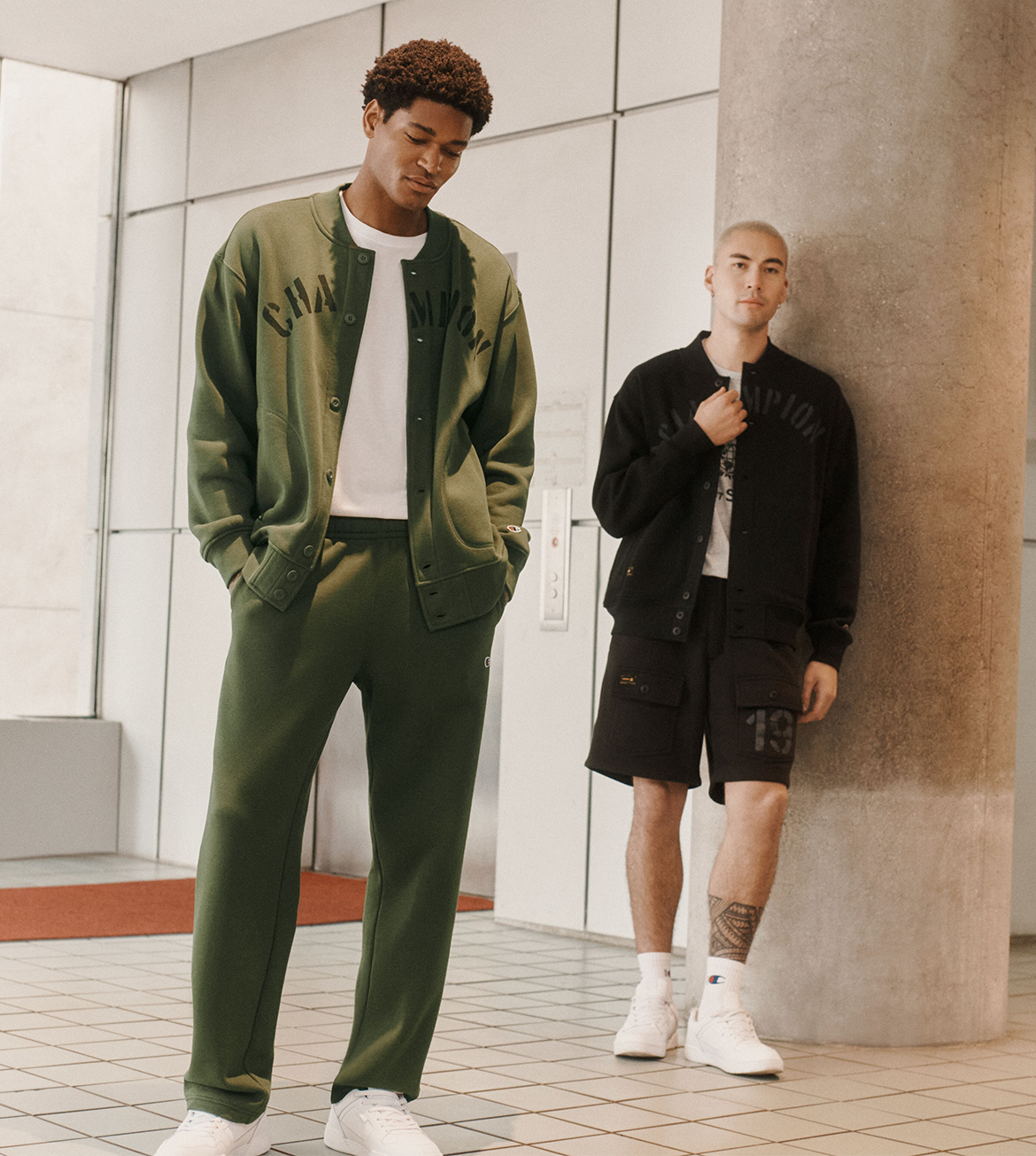 Two male models wearing streetwear apparel in a modern indoor setting with tiled floors and a large concrete column. The model on the left wears an olive green varsity jacket and matching pants over a white t-shirt, while the model on the right wears a black varsity jacket with matching black shorts over a white t-shirt.