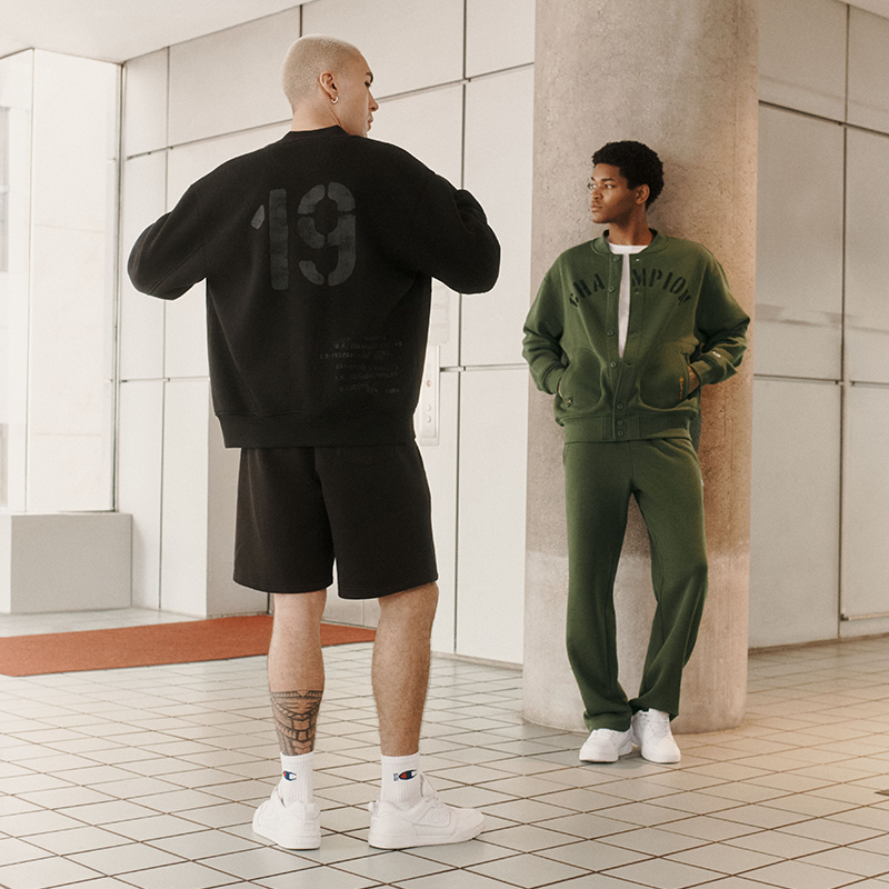 Two models in an indoor modern setting, one showing the back of a black sweatshirt with a large '19' graphic and matching black shorts, while the other models a green zip-up sweatshirt and matching green sweatpants.