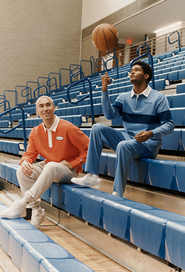 Two men sitting on blue gymnasium bleachers, one wearing an orange polo shirt and light pants, the other wearing a blue striped sweater and matching pants, with one man balancing a basketball on his finger.
