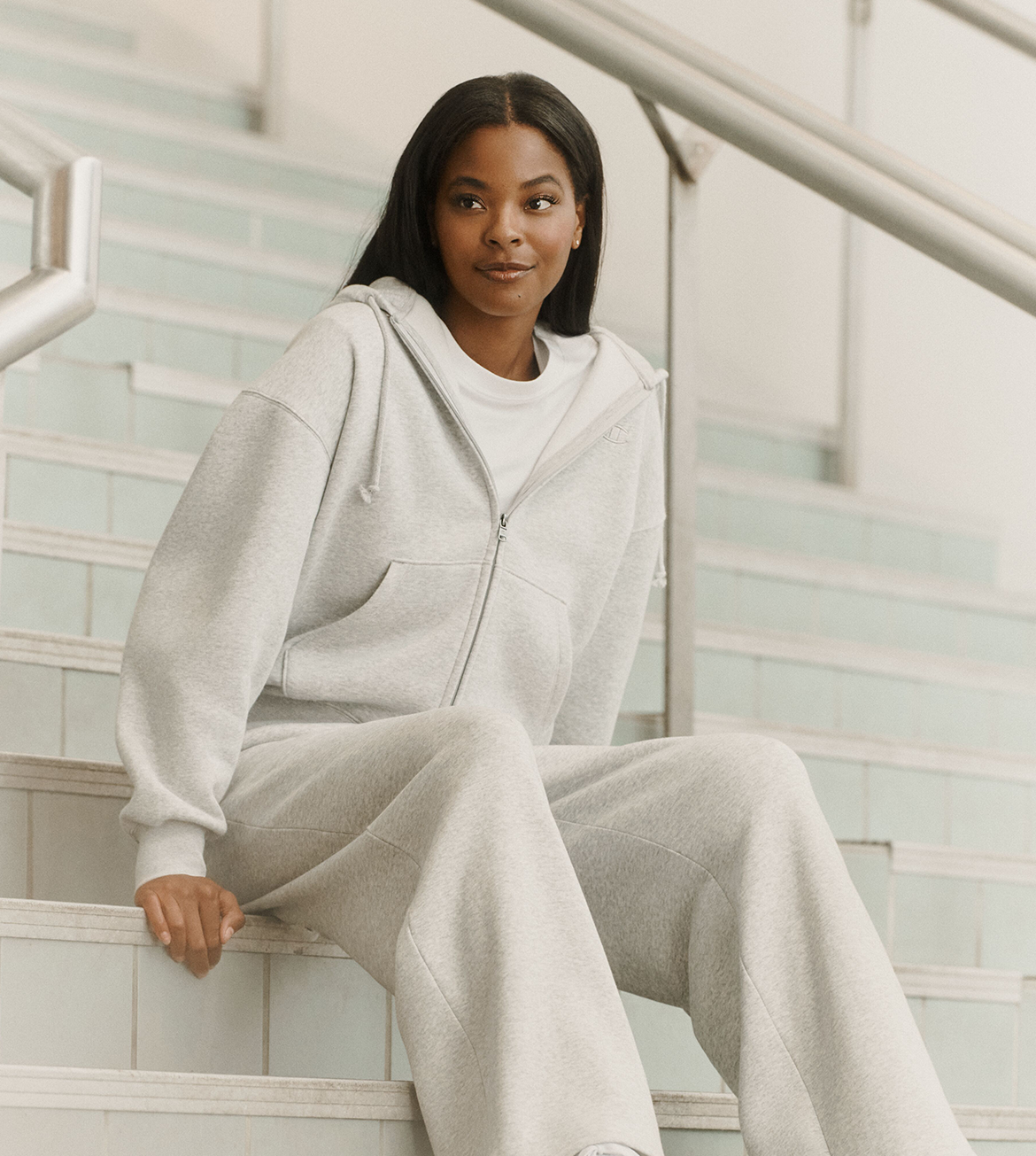 A woman sits on indoor stairs wearing a light heather gray zip-up hoodie and matching sweatpants over a white top.