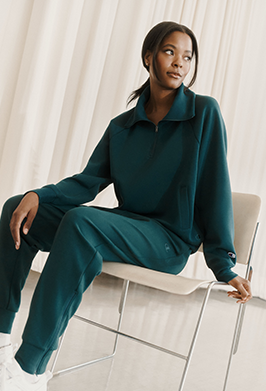 A woman wearing a matching dark teal green lounge set consisting of a quarter-zip long-sleeve top and jogger pants, sitting on a modern white chair with a metal frame against a backdrop of soft white curtains.