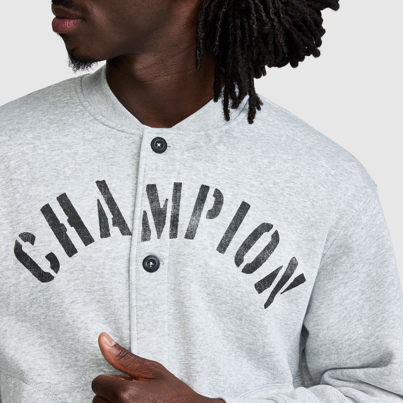 A person wearing a grey Champion Fleece Baseball Jacket with the word CHAMPION printed in large black letters across the chest.