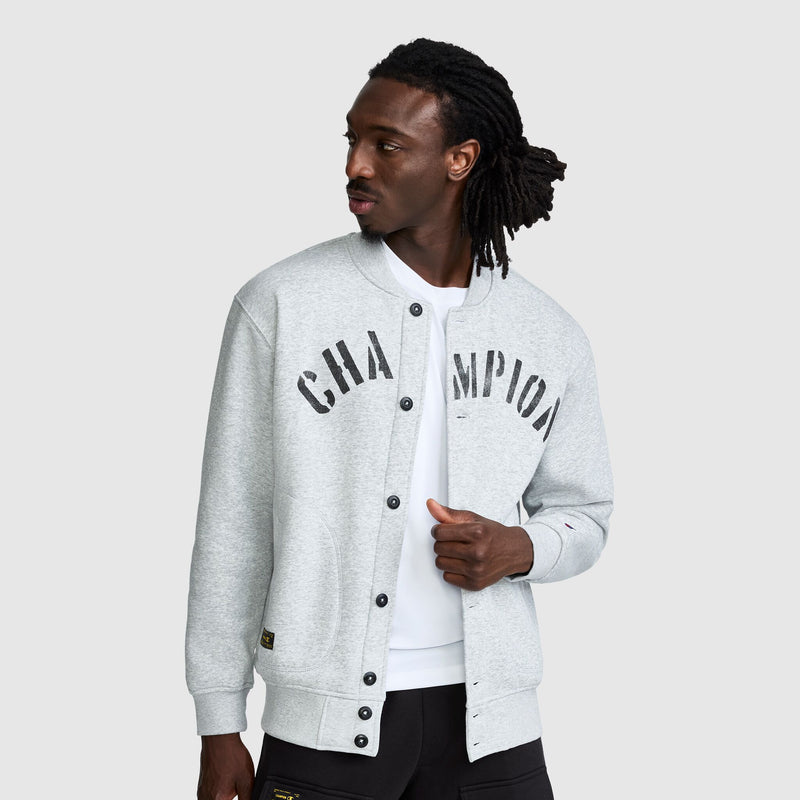 A person wearing a grey Champion Fleece Baseball Jacket with a distressed black logo on the chest.