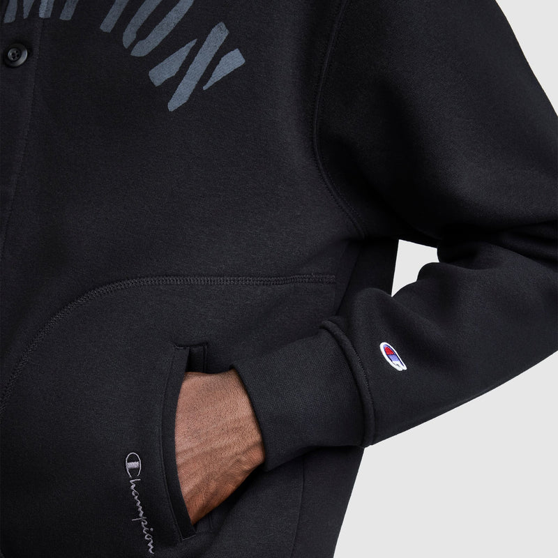 A person is wearing a black Champion Fleece Baseball Jacket, casually placing one hand in the pocket. The Champion logo appears on the cuff and near the pocket, adding a vintage athletic touch to this classic varsity jacket style. The background is plain and light-colored.