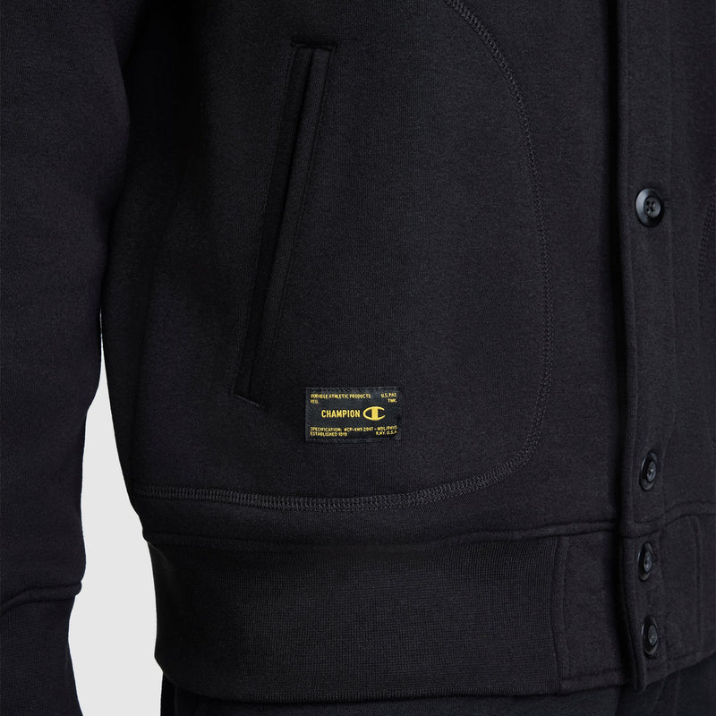 Close-up of a person wearing the Champion Fleece Baseball Jacket in black, featuring side pockets, button closure, and a small yellow Champion logo tag above the waistband on the left side. This varsity jacket showcases vintage athletic style inspired by classic designs from Champion.