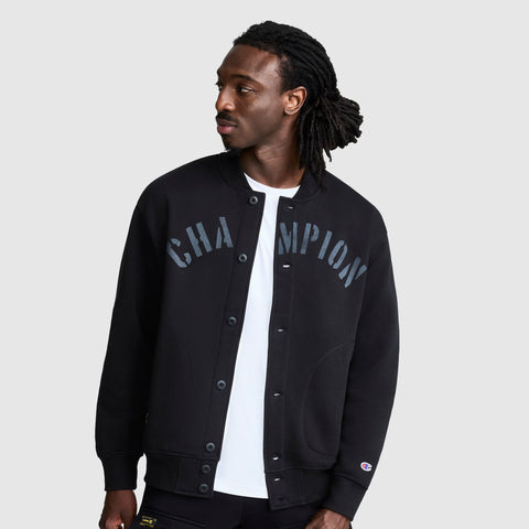 A man with long dreadlocks wears the Champion Fleece Baseball Jacket in black over a white shirt, looking to his left. The vintage athletic style of this varsity jacket stands out against the plain light gray background.