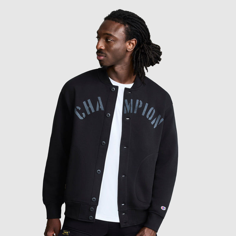 A man with long dreadlocks wears the Champion Fleece Baseball Jacket in black over a white shirt, looking to his left. The vintage athletic style of this varsity jacket stands out against the plain light gray background.